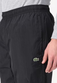 Pantaloni negri ușori cu un talie elastică, buzunare laterale și un logo brodat cu crocodil verde aproape de buzunarul din dreapta. Textură moale.