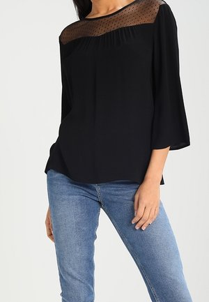 Haut noir avec encolure transparente à pois, coupe décontractée, manches trois-quarts et tissu lisse. Associé à un jean en denim bleu clair.