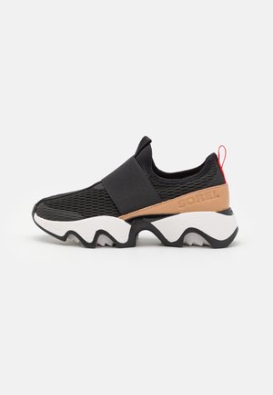 Czarny sneaker wsuwany z siateczkową cholewką, szerokim elastycznym paskiem, beżowym paskiem na pięcie z logo Sorel, czerwoną pętelką do zakładania i masywną białą i czarną podeszwą.