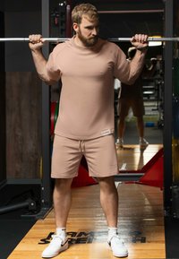 Tenue de sport brun clair comprenant un haut à manches courtes et un short. Le matériau semble doux avec un branding minimaliste. Des baskets blanches sont incluses.