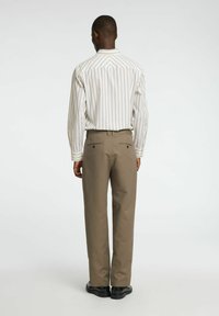 Widok z tyłu osoby noszącej białą, długą koszulę w pionowe paski oraz beżowe spodnie, wyposażone w kieszenie i o gładkiej teksturze.