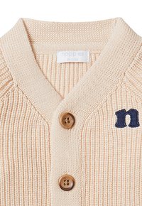 Beige gestrickte Strickjacke mit V-Ausschnitt, ausgestattet mit zwei braunen Knöpfen und einem navyblau gestickten Buchstaben 'n' auf der linken Seite.