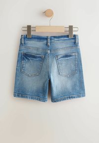 Shorts en denim bleu clair, présentant un design classique à cinq poches, des bords effilochés et un léger délavage. Suspendus sur un cintre en bois.