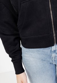 Czarna bluza rozpinana z prążkowanymi mankietami i u dołu, posiadająca przednią kieszeń i miękką teksturę, zestawiona z jasnoniebieskimi spodniami typu high-waisted.