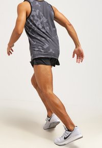 Homme portant un débardeur camouflé noir, un short noir et des chaussures de course Nike blanches, en plein mouvement sur un fond uni.