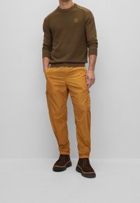 Brun ribbad långärmad tröja, gula cargobyxor med fickor och bruna snörade kängor med tjocka sålar. Neutral bakgrund.