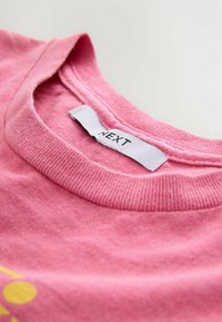 Pink cotton T-shirt with a ribbed crew neckline. Features a label reading "NEXT." Texture appears soft and smooth.