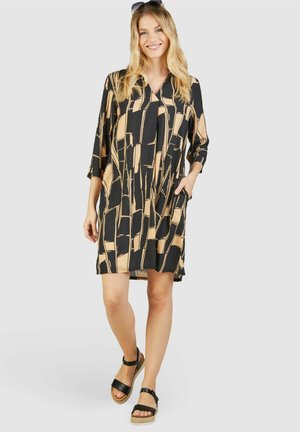 Schwarz- und braun gemustertes Kleid mit Dreiviertelärmeln, einem V-Ausschnitt und Taschen, kombiniert mit schwarzen Sandalen mit einem blockigen Absatz.