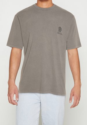 Mann trägt ein mattbraunes T-Shirt mit kurzen Ärmeln und kleinem dunklen Logo auf der linken Brustseite sowie hellblaue Jeans vor neutralem Hintergrund.