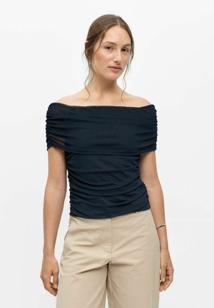 Femme à la peau claire et aux cheveux bruns porte un haut noir froncé sans épaules et un pantalon beige sur un fond blanc uni.