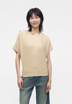 Person står og smiler, iført en kortærmet beige top med runde knapper ved halsudskæringen og blå jeans på en ensfarvet, lys baggrund.