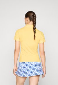 Polo shirt giallo a coste con maniche corte e dettagli bianchi, abbinato a una gonna blu con disegni circolari.
