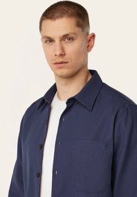 Marineblauwe knoopshirt met een textuurstof, een kraag en een linkerborstzak. Bevat zwarte knopen en een tonale logo op de zak.