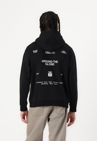 Schwarzer Kapuzenpullover mit einem großen Grafikdesign auf dem Rücken, das "AROUND THE GLOBE" und verschiedene Städtenamen in weißem Text enthält. Einfache Vorderseite.