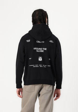 Schwarzer Kapuzenpullover mit einem großen Grafikdesign auf dem Rücken, das "AROUND THE GLOBE" und verschiedene Städtenamen in weißem Text enthält. Einfache Vorderseite.
