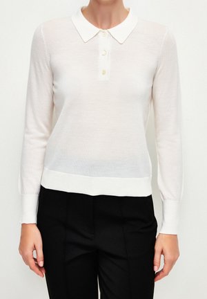 Kobieta nosząca biały, długi sweter polo z trzema guzikami, zestawiony z czarnymi, dopasowanymi spodniami, pokazana od szyi do bioder.