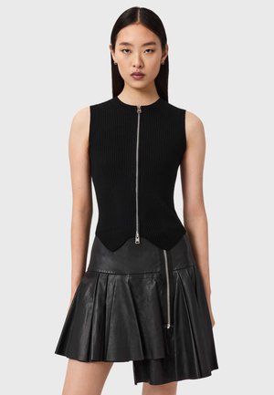 Nainen pukeutuneena mustaan hihattomaan resoripaitaan, jossa on edessä vetoketju, sekä epäsymmetriseen mustaan nahkaan, jossa on laskostettu hame ja vetoketju.