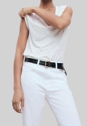 Osoba nosząca biały top bez rękawów i białe spodnie, poprawiająca lewy ramię topu prawą ręką, widoczny czarny pasek.