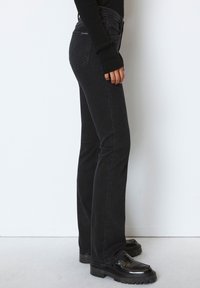 Jean noir à coupe droite avec une texture lisse, doté de poches arrière et d'une taille mi-haute, associé à des chaussures noires imposantes.