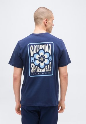 Uomo con testa rasata che indossa una maglietta blu navy Columbia Sportswear con logo floreale retrò e testo sul retro, in piedi davanti a uno sfondo semplice.