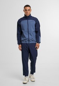 Vīriešu sporta apģērbs, kurā ir zils un tumši zils vējjaka ar rāvējslēdzēju priekšā un elastīgu jostasvietu, apvienots ar saskaņotām tumši zilām biksēm un baltām sporta apaviem.