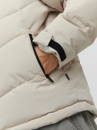 Beige Daunenjacke, die ein Handgelenk mit verstellbarem Bündchen und einer mit einem schwarzen Rand versehenen Reißverschlusstasche zeigt. Glatte Textur mit gestepptem Design.