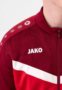 Rote Sportjacke mit weißen Akzenten, strukturiertem Stoff, Reißverschluss und Logo. Besitzt ein gemustertes Design und kreisförmige Schulterdetails.
