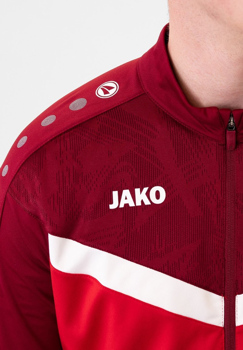 Rote Sportjacke mit weißen Akzenten, strukturiertem Stoff, Reißverschluss und Logo. Besitzt ein gemustertes Design und kreisförmige Schulterdetails.