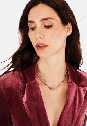 Hopeinen kaulakoru, jossa pieniä pyöreitä lenkkejä ja keskellä pitkäomainen korostus, puettuna syvän viininpunaiseen samettitakkiin, jossa on kärkipäiset kaulukset.