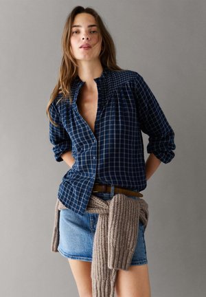 Vrouw met lang bruin haar draagt een blauw geruit hemd, een denim rok, een bruine riem en een beige gebreide trui gestrikt rond haar taille, staand tegen een grijze muur.