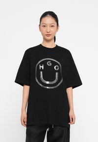 Camiseta negra de gran tamaño hecha de algodón, con un diseño de logo de purpurina plateada en un patrón circular en la parte frontal. Mangas cortas.