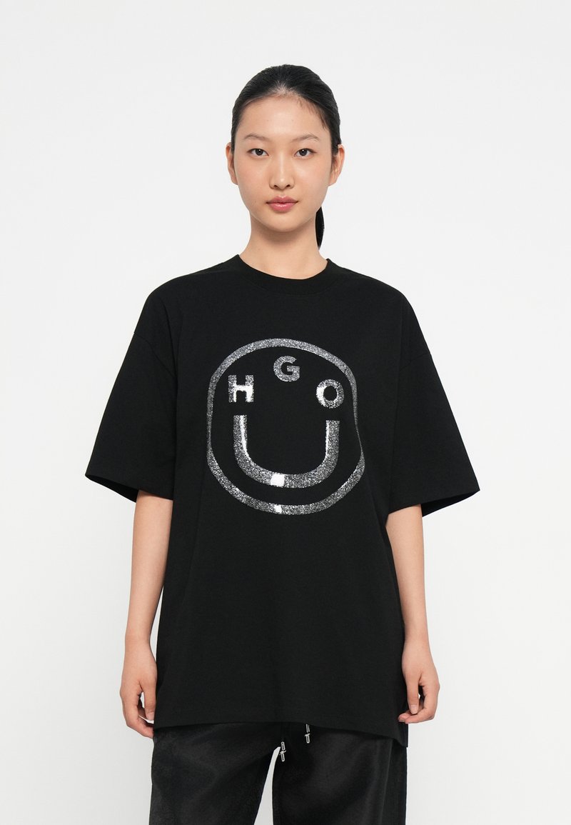 Camiseta negra de gran tamaño hecha de algodón, con un diseño de logo de purpurina plateada en un patrón circular en la parte frontal. Mangas cortas.