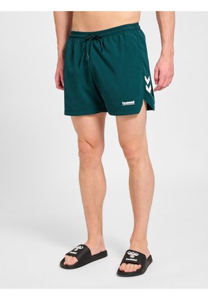 Person står iført turkis sports-shorts med hvidt chevron-mønster og sorte Hummel-slippers på en ensfarvet lys baggrund.