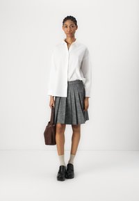 Hvit langermet skjorte med knapper, grå rutete plissébelte, brun skulderveske, beige sokker, svarte sko, minimalistisk bakgrunn.