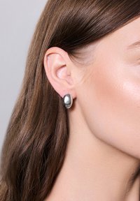 Srebrny okrągły wkręt z gładką, refleksyjną powierzchnią, umieszczony na uchu modelki, w towarzystwie prostych, brązowych włosów.