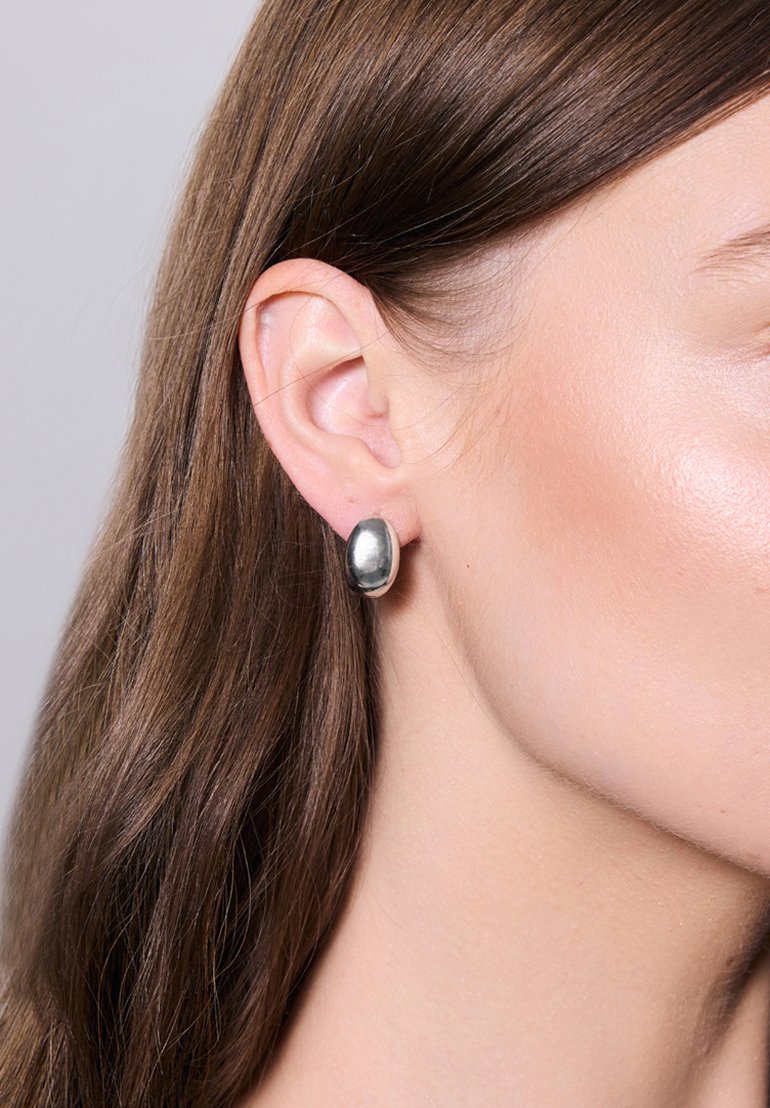 Srebrny okrągły wkręt z gładką, refleksyjną powierzchnią, umieszczony na uchu modelki, w towarzystwie prostych, brązowych włosów.