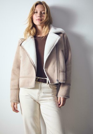 Blonde vrouw in een beige schapenleren jack, een bruine top, crèmekleurige broek en een bruine riem, staand tegen een eenvoudige witte muur.