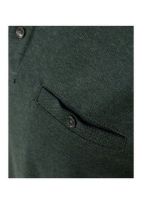 Dunkelgrünes, strukturiertes Poloshirt mit einem knöpfbaren Kragen und einer kleinen Brusttasche, die mit einem runden schwarzen Knopf versehen ist.