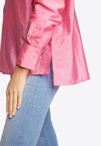 Rosa Satinhemd mit langen Ärmeln und geknöpften Manschetten, ausgestattet mit einem Seitenschlitz und glatter Textur. Kombiniert mit hellblauen Jeans.