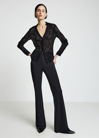 Schwarze Spitzenbluse mit durchgehender Knopfleiste, langen Ärmeln und V-Ausschnitt. Kombiniert mit hoch taillierten, schwarzen ausgestellten Hosen und spitzen schwarzen Schuhen.