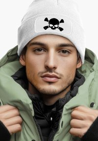 White knit beanie with a black skull and crossbones patch, worn with an olive green puffy jacket and a black inner lining.