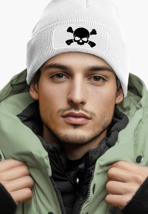 White knit beanie with a black skull and crossbones patch, worn with an olive green puffy jacket and a black inner lining.