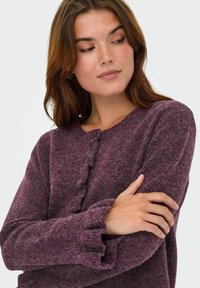 Cardigan en laine violet avec un col rond, fermeture à boutons et manchettes à volants. Texture douce avec un aspect tricoté et confortable.