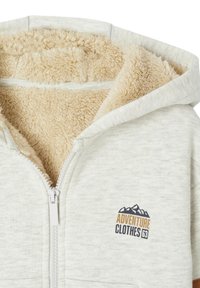 Light gray zip-up hoodie with a beige fleece lining. Features an embroidered logo with mountains and text "ADVENTURE CLOTHES 63." Smooth texture.
