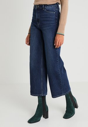 Persona que lleva vaqueros de tiro alto y pierna ancha de color azul oscuro, camiseta beige de manga larga y botines verdes oscuros de ante con tacones gruesos.