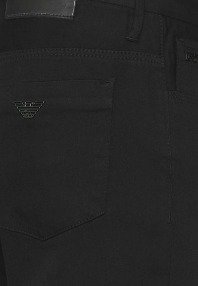 Jeans en denim noir arborant un logo d'aigle distinctif sur la poche arrière, avec des coutures et une texture lisse et mate.