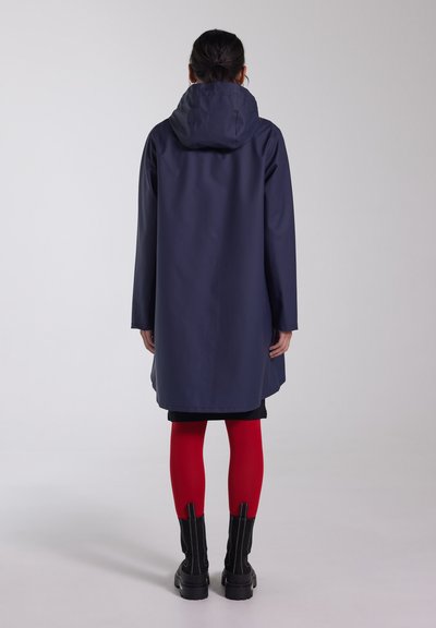Marine waterdichte jas met een capuchon, lange mouwen en een rechte snit, gecombineerd met rode leggings en zwarte enkellaarsjes, gezien vanuit de achterkant.