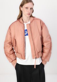 Pink bomber jacket with a smooth texture, ribbed cuffs, and a zipper front. Worn over a white shirt with a blue logo, paired with black pants.
