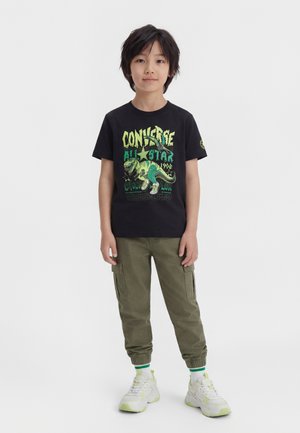 Dreng iført sort Converse All Star T-shirt med grønt dinosaurprint, olivengrøn cargo-buks, hvide sneakers med grønne detaljer på hvid baggrund.