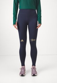 Marinblå leggings med hög midja, prydda med guld Adidas-logotyp och texten "50 BM Marathon". Slät, elastisk tyg, tillsammans med ljuslila sneakers.
